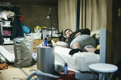 散らかった部屋で肘枕をして寝転がるデブ男性