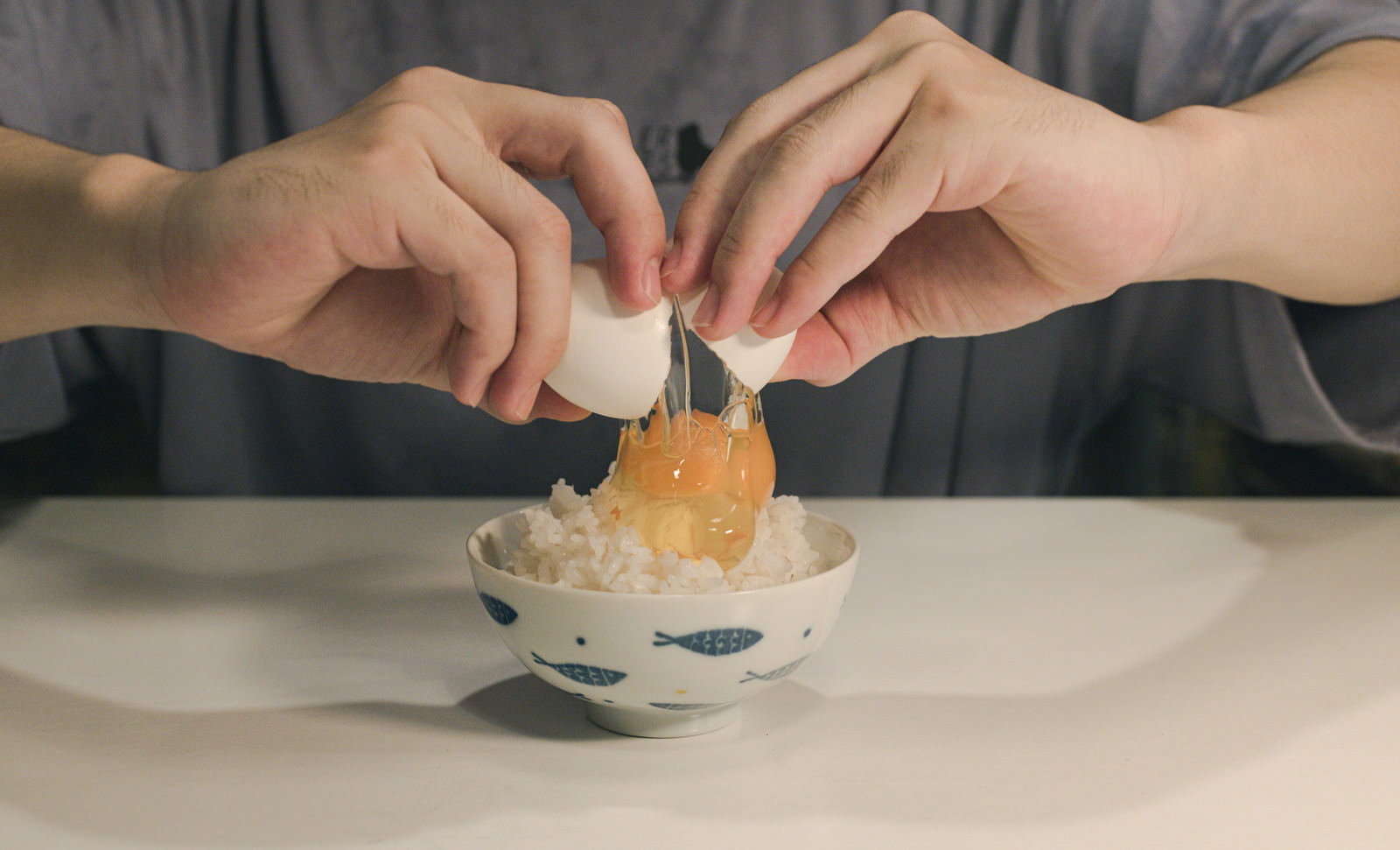 白い茶碗のご飯の上で両手で卵の殻を割って卵かけご飯を作る様子