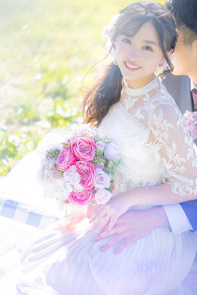 ピンクのバラのブーケを持つ白いウェディングドレス姿の花嫁が笑顔で新郎に寄り添う［モデル：河村友歌］