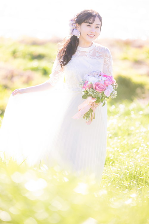 緑の野原で白いドレス姿の花嫁がピンクのブーケを手に微笑む