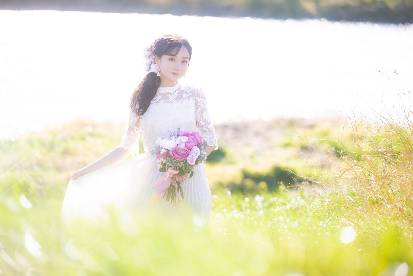 春の野原でピンク色のブーケを持つ白いウェディングドレスの花嫁［モデル：河村友歌］