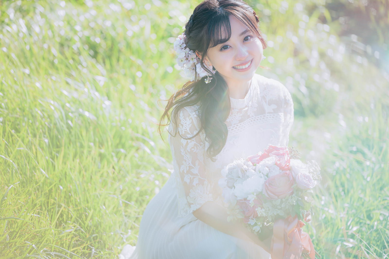白いウエディングドレスを着た花嫁が、ピンクと白のブーケを持ち、緑の屋外で笑顔でポーズを取っている［モデル：河村友歌］