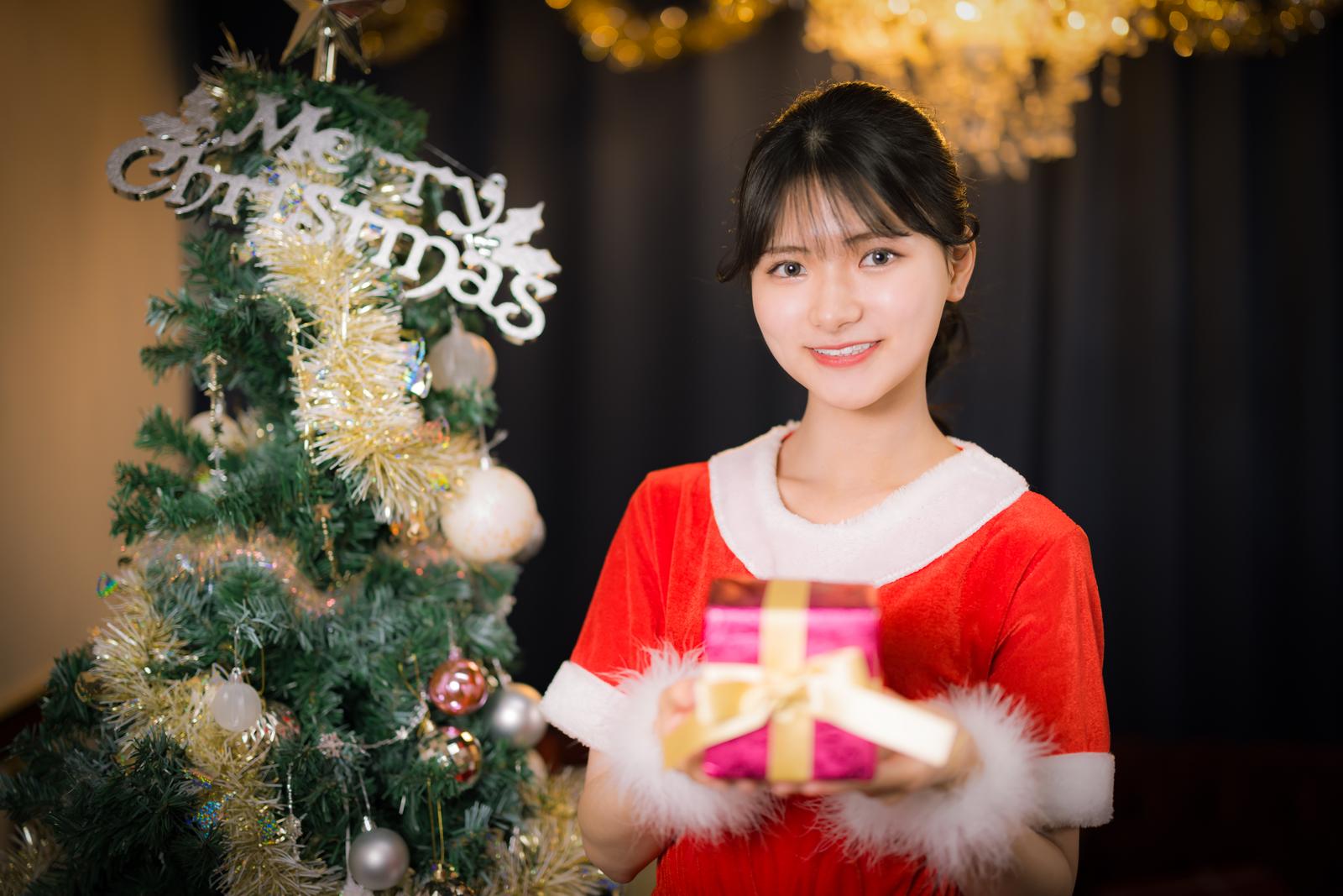 クリスマスツリーの前でプレゼントを持つサンタコス姿の女性［モデル：モデルリリース］