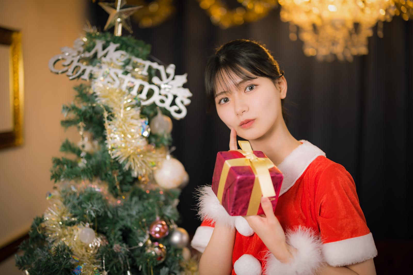 サンタコス姿の女性がゴールドリボンの赤いプレゼントボックスを持ち、クリスマスツリーの前に立っている