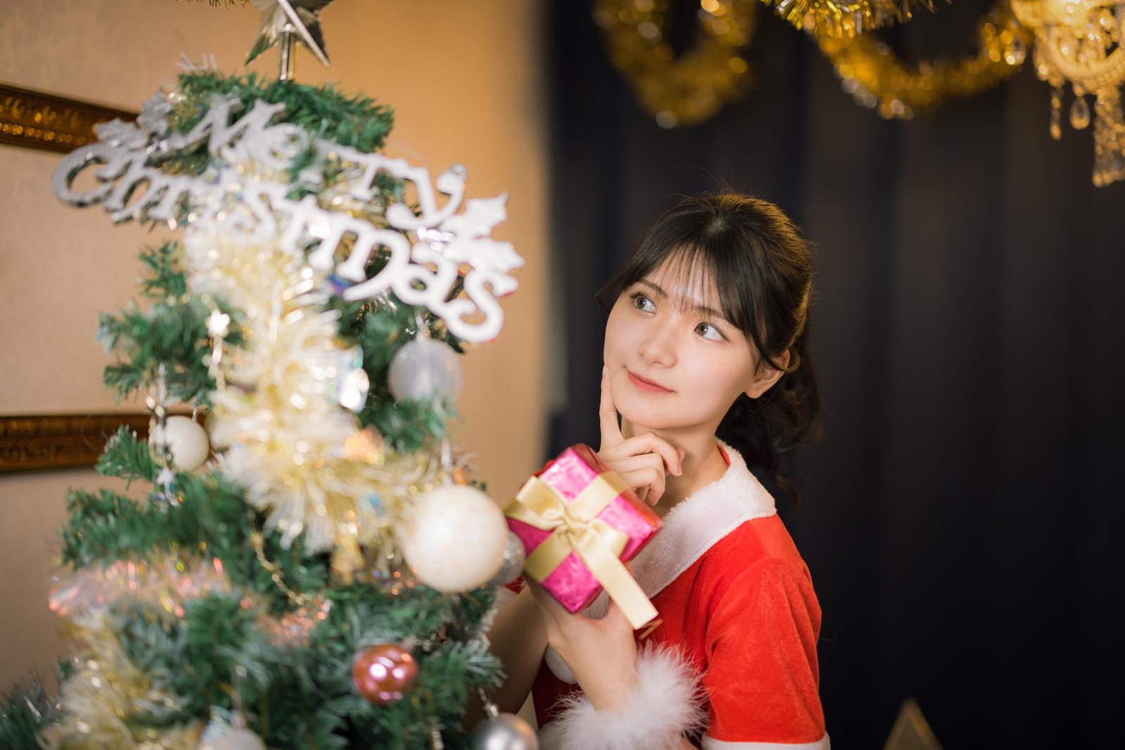 赤いサンタコス姿の女性がプレゼント箱を持ってクリスマスツリーを見上げている［モデル：モデルリリース］