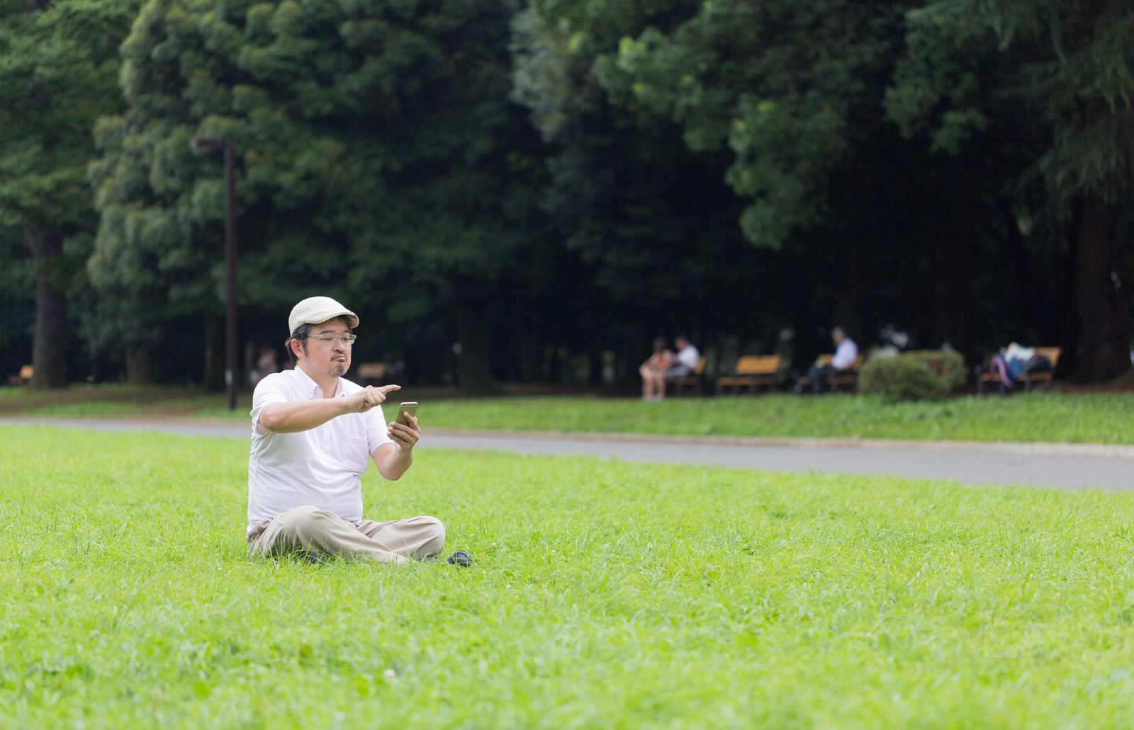 公園の芝生に座ってスマホを操作するハンチング帽の男性［モデル：ナリシゲ］