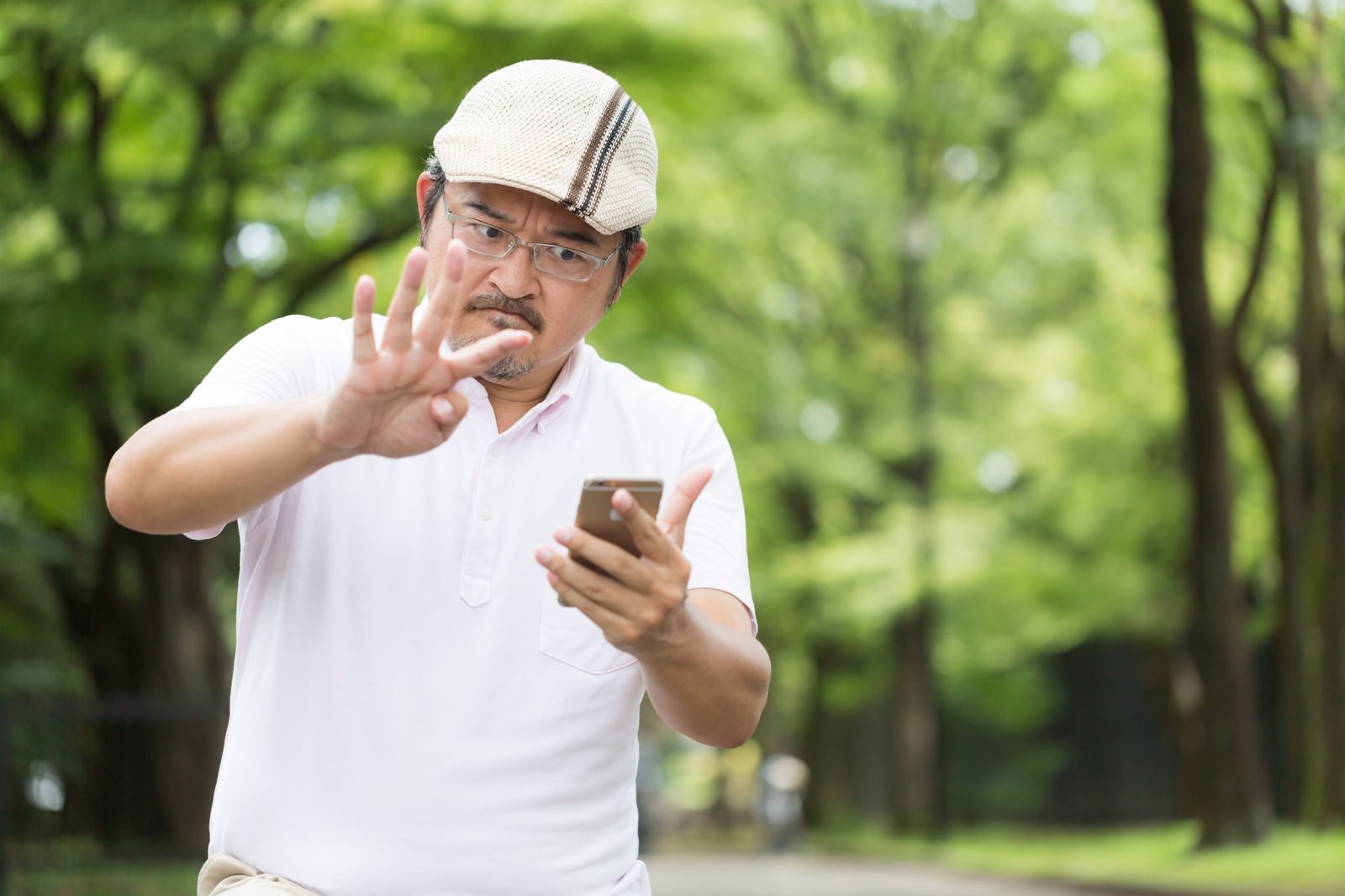 公園でスマートフォンを見ながら捕まえるジェスチャーをする眼鏡をかけた男性［モデル：ナリシゲ］
