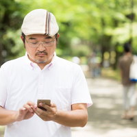 家族連れが多い公園で歩きスマホをするハンチング帽の男性の写真