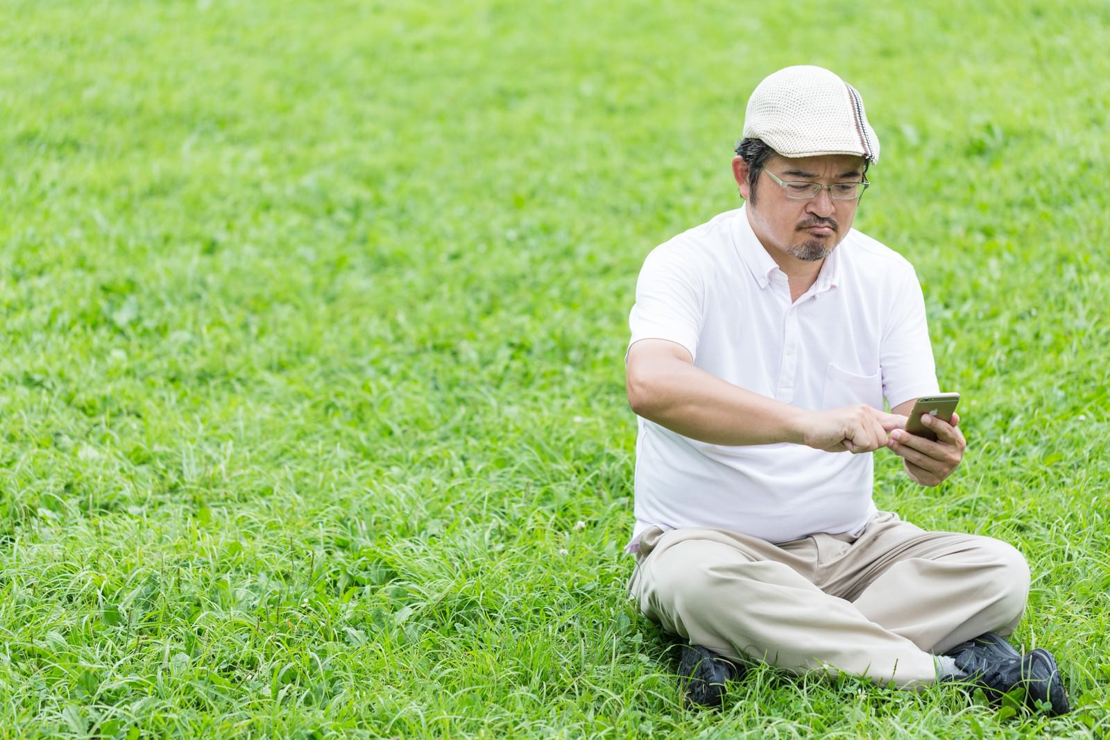 白いハンチング帽をかぶった男性が、公園の芝生に座ってスマートフォンを両手で握り操作している［モデル：ナリシゲ］
