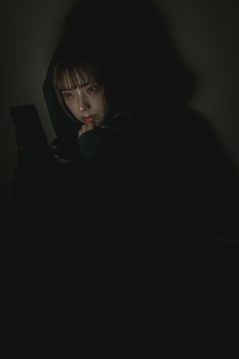 暗がりでスマートフォンの画面を見つめている女性［モデル：高尾実生］