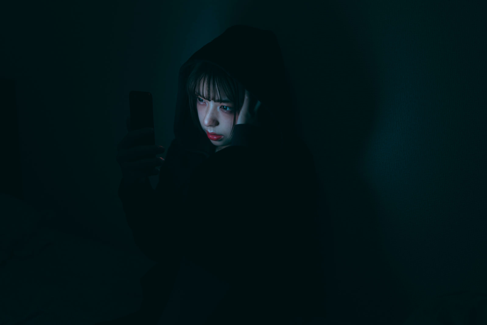暗い部屋でスマートフォンの画面を見つめる女性の様子［モデル：高尾実生］