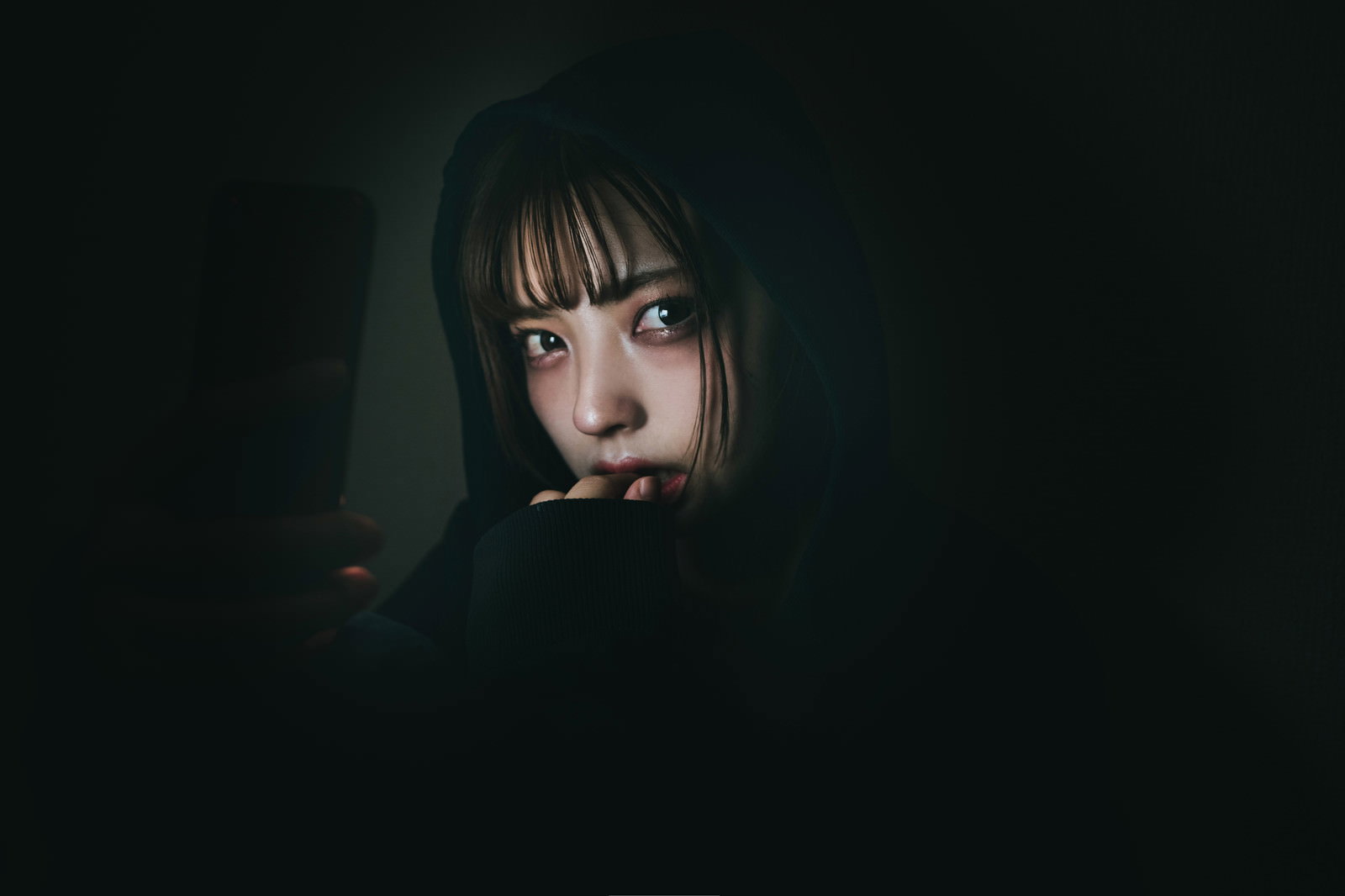 暗がりでスマートフォンを見つめる女性の表情［モデル：高尾実生］