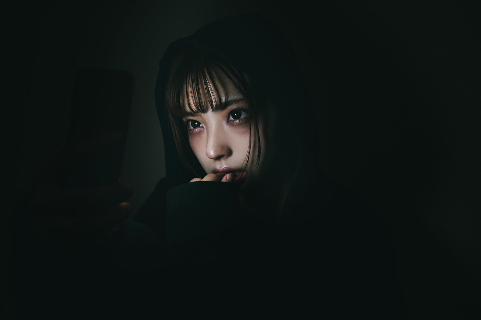 暗い場所でスマートフォンの画面を見つめる女性［モデル：高尾実生］
