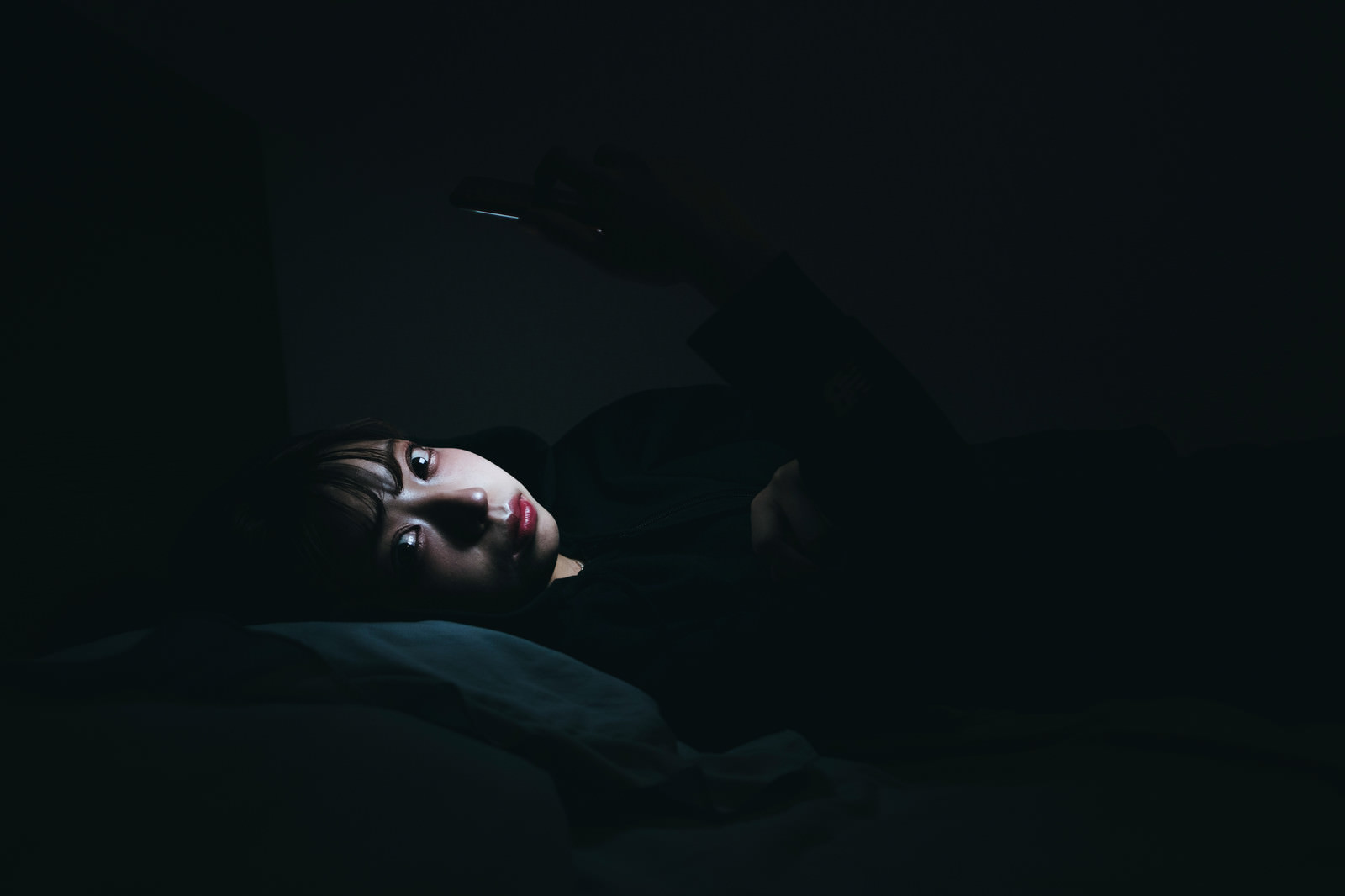暗い部屋でスマートフォンの画面を見つめる女性［モデル：高尾実生］