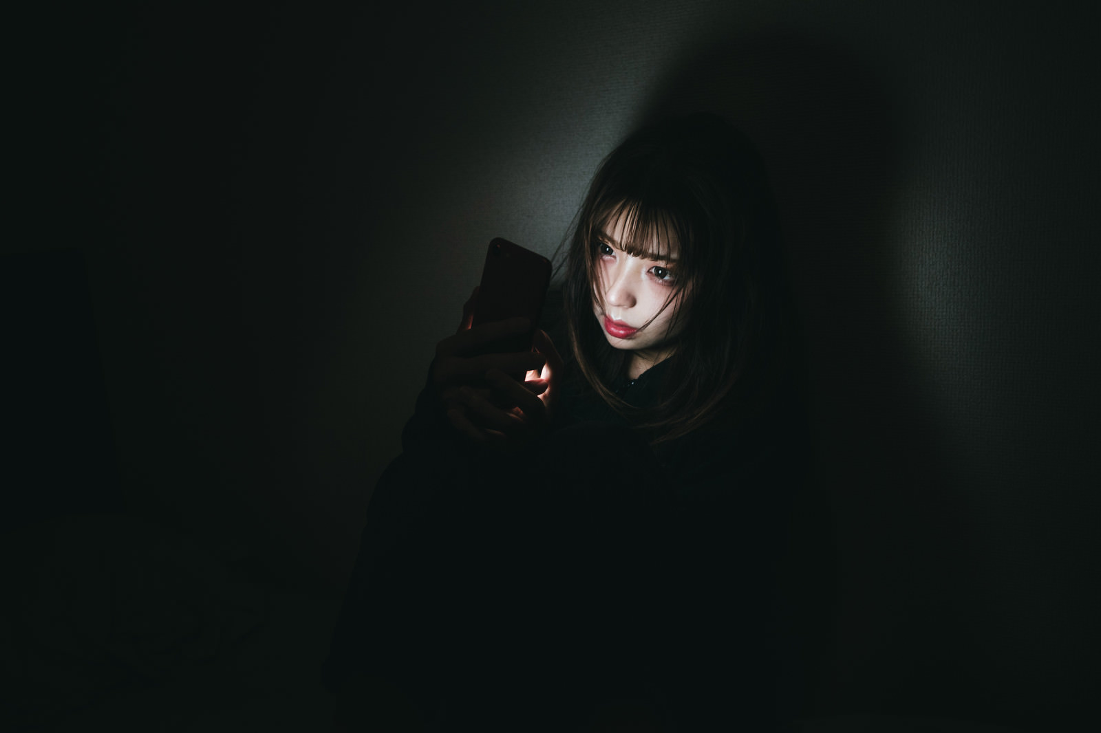 暗い室内でスマートフォンを見つめる女性［モデル：高尾実生］