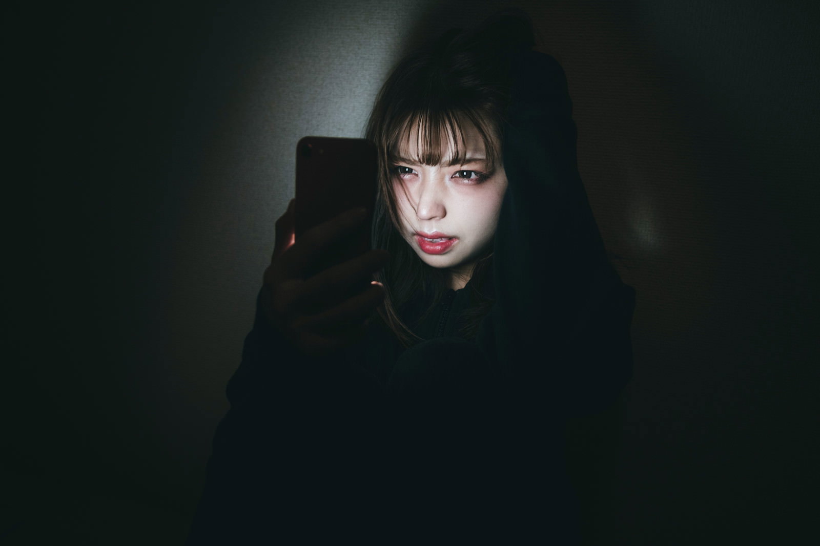 暗い場所でスマートフォンを見つめる不機嫌そうな女性［モデル：高尾実生］