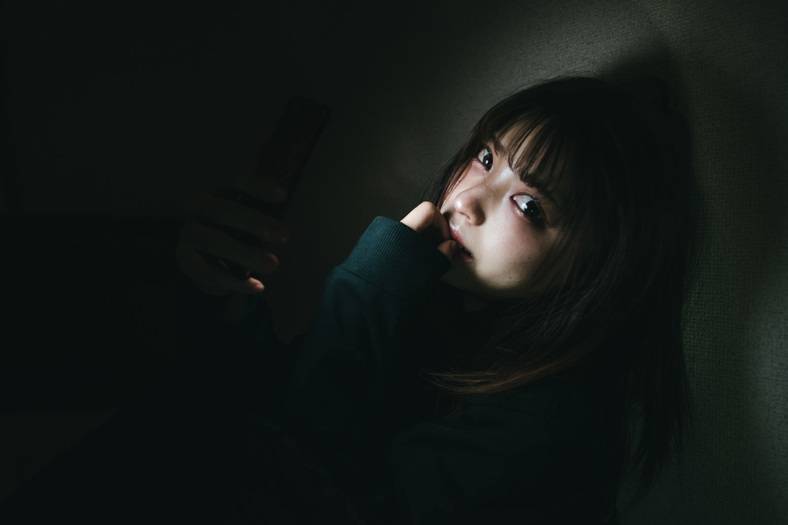 暗い部屋で手で頭を支えながらスマホを見つめる女性［モデル：高尾実生］