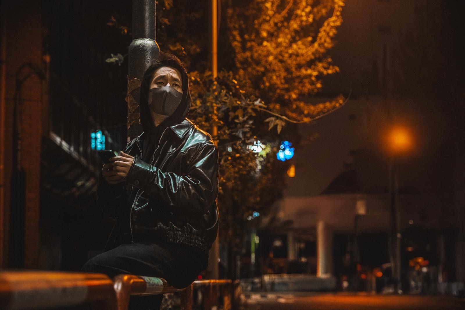 夜の街のベンチでフードとマスク姿でスマートフォンを握る男性［モデル：大川竜弥］