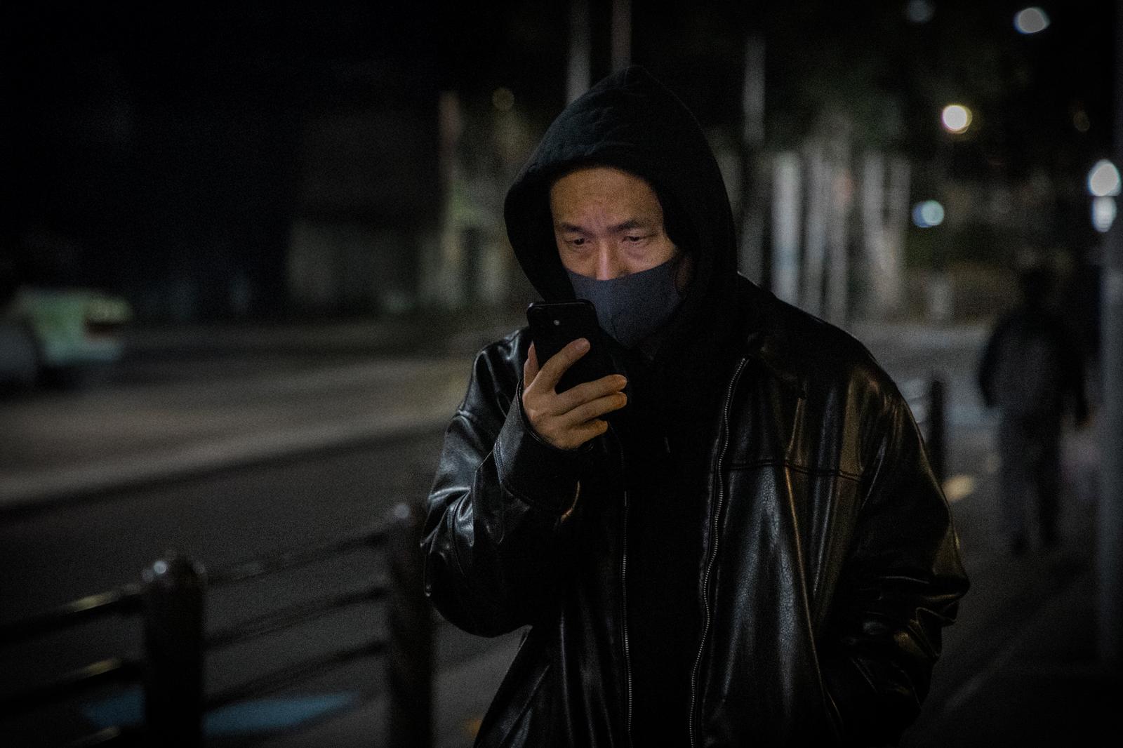 黒いレザージャケットとフードを被り、マスクで顔を覆った男性がスマートフォンを持ちながら夜間の路上に立っている［モデル：大川竜弥］