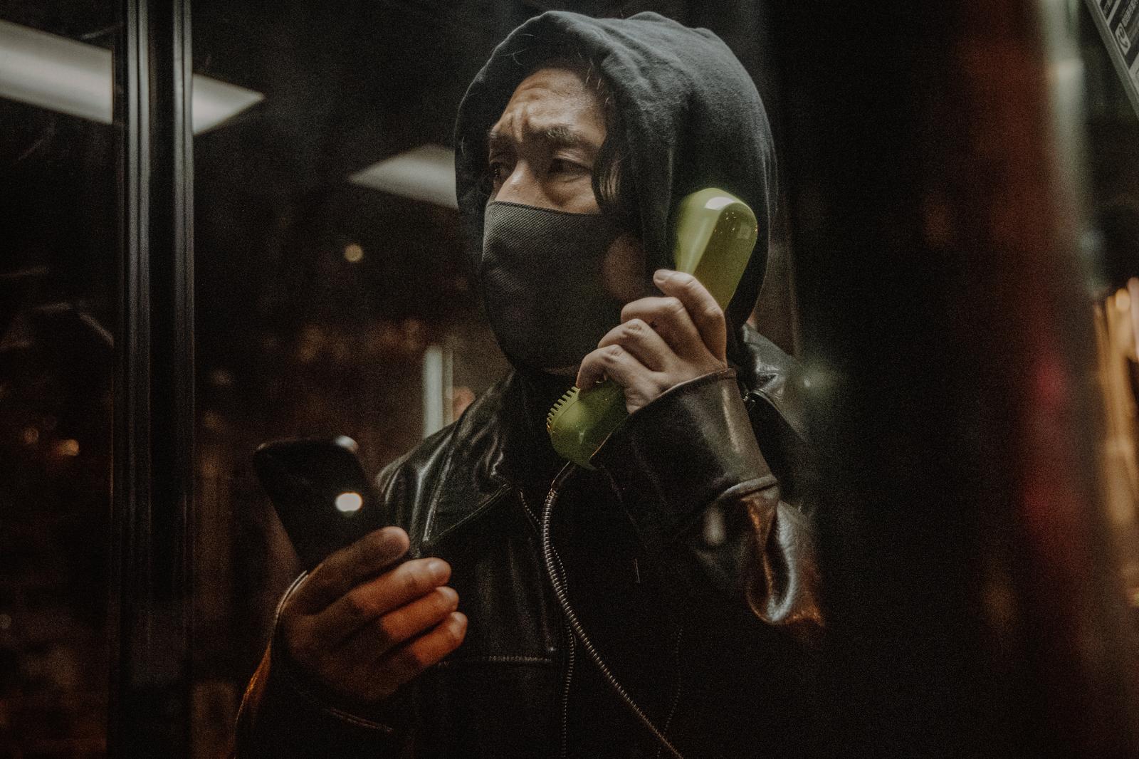 公衆電話ボックスで通話する黒いジャケット姿の男性［モデル：大川竜弥］