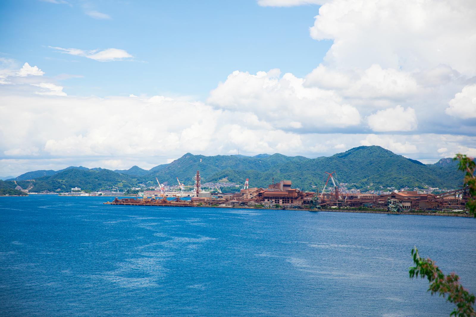 広島の海峡の向こうに見える工場とクレーン、背景に緑の山々と青空