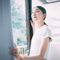 白いTシャツ姿でカーテンを開けて朝日を浴びる女性の横顔の写真