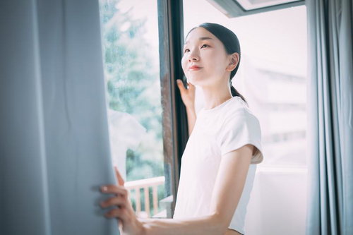 白いTシャツ姿でカーテンを開けて朝日を浴びる女性の横顔