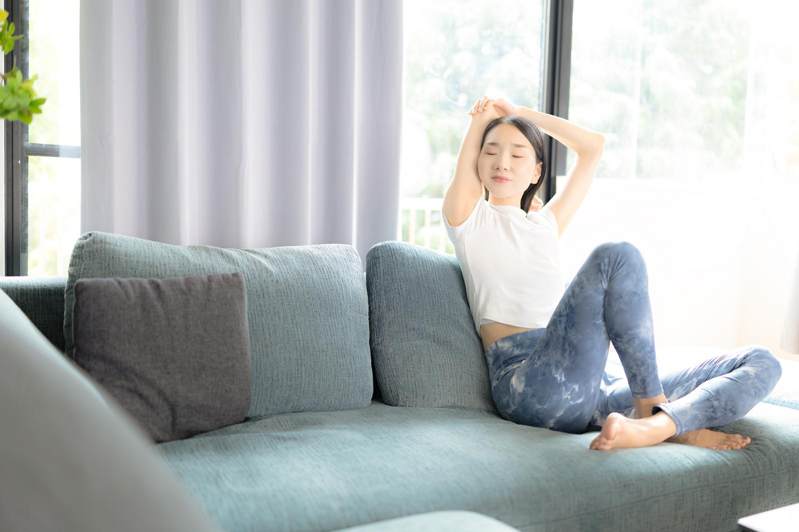 グレーソファに座り両手を上げてストレッチをする白Tシャツの女性［モデル：ひるかわえみこ］