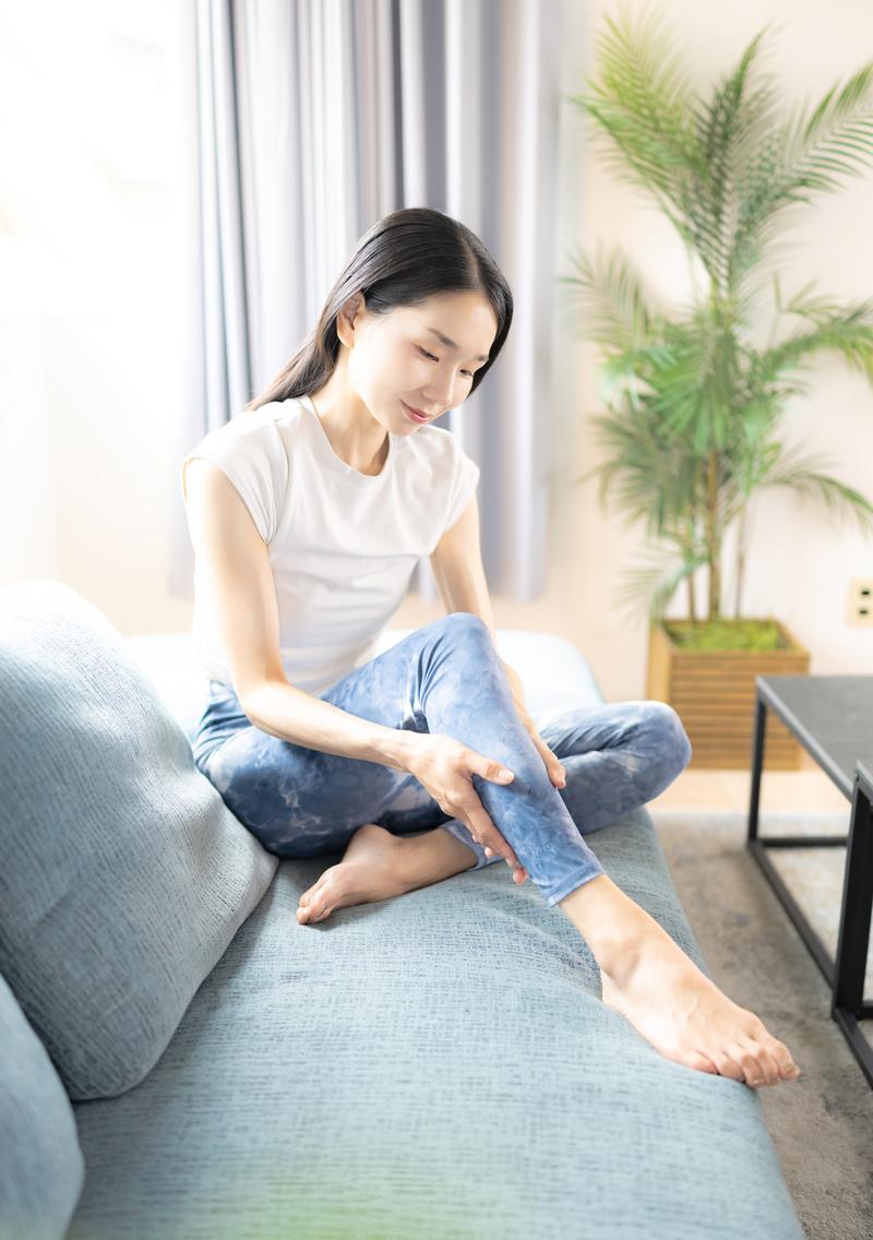グレーのソファに座ってふくらはぎをマッサージしている女性［モデル：ひるかわえみこ］