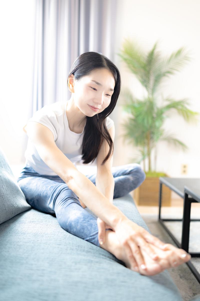 グレーのソファに座って脚を伸ばしストレッチをする女性［モデル：ひるかわえみこ］