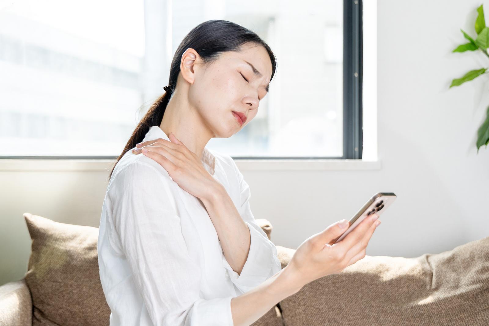 ソファに座りスマホを見ながら首の痛みを感じて手を当てる女性［モデル：ひるかわえみこ］