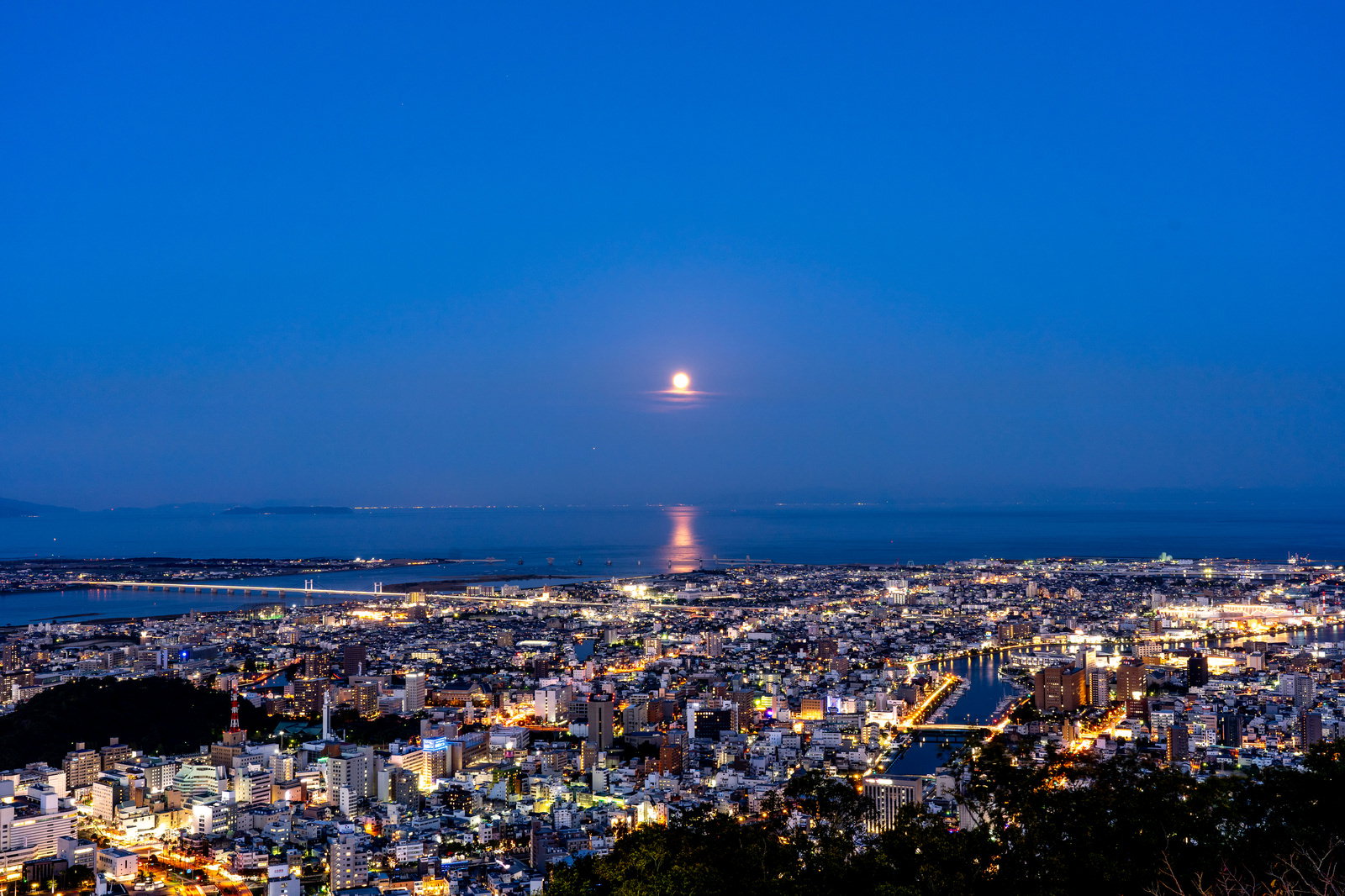青い夜空に満月が浮かび、眼下に徳島市街地の夜景が広がる風景