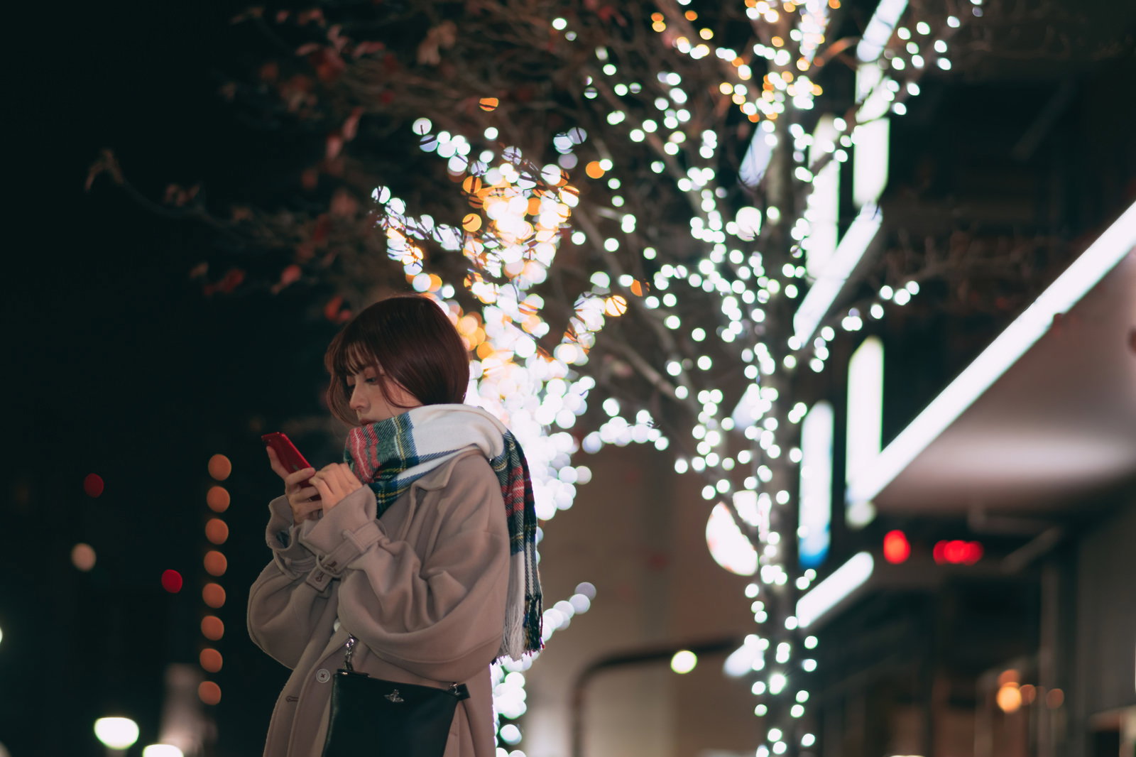 イルミネーションが輝く夜の街でスマートフォンを見る女性［モデル：高尾実生］