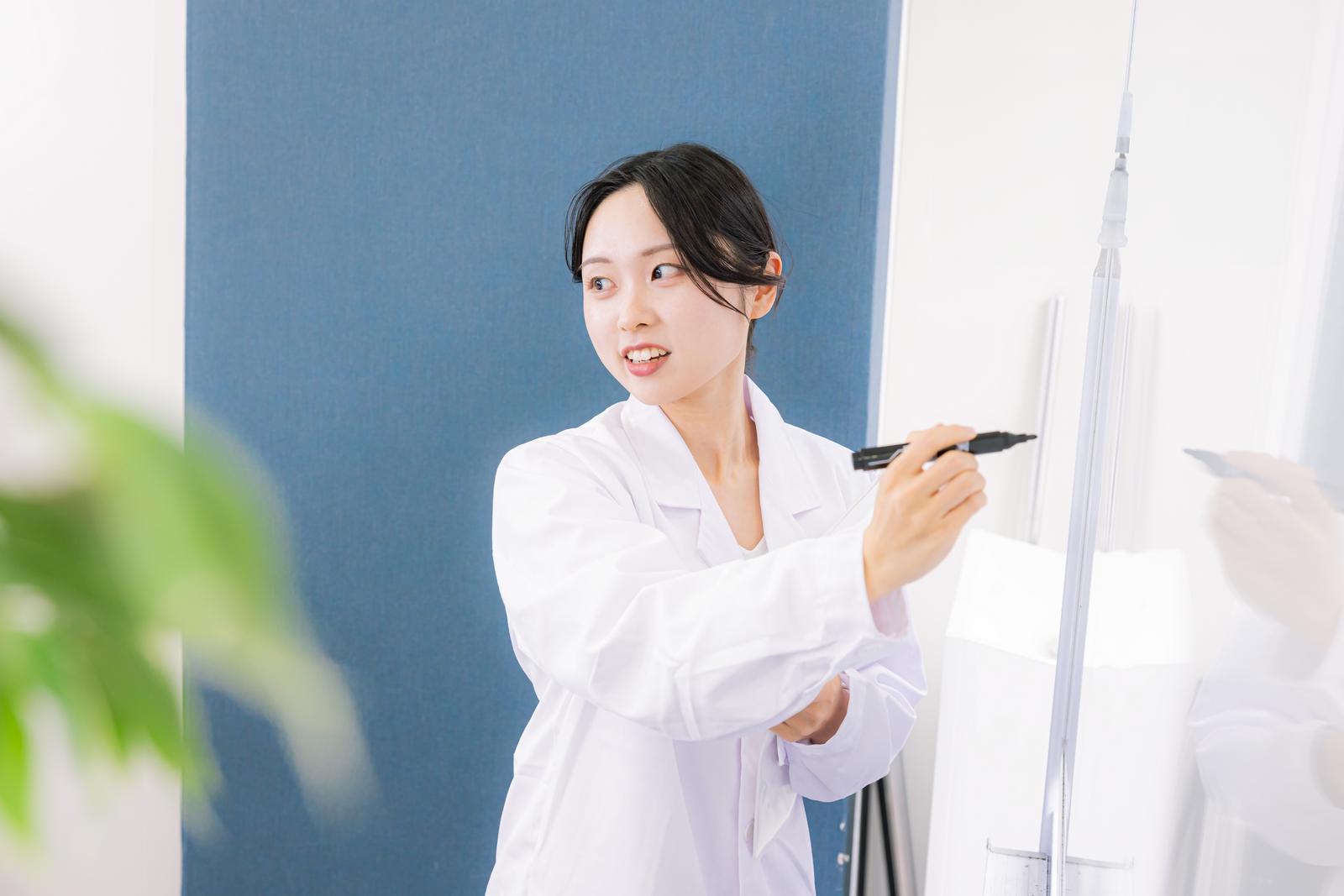 ホワイトボードにマーカーで書きながら説明する白衣姿の女性［モデル：吉屋ゆり］