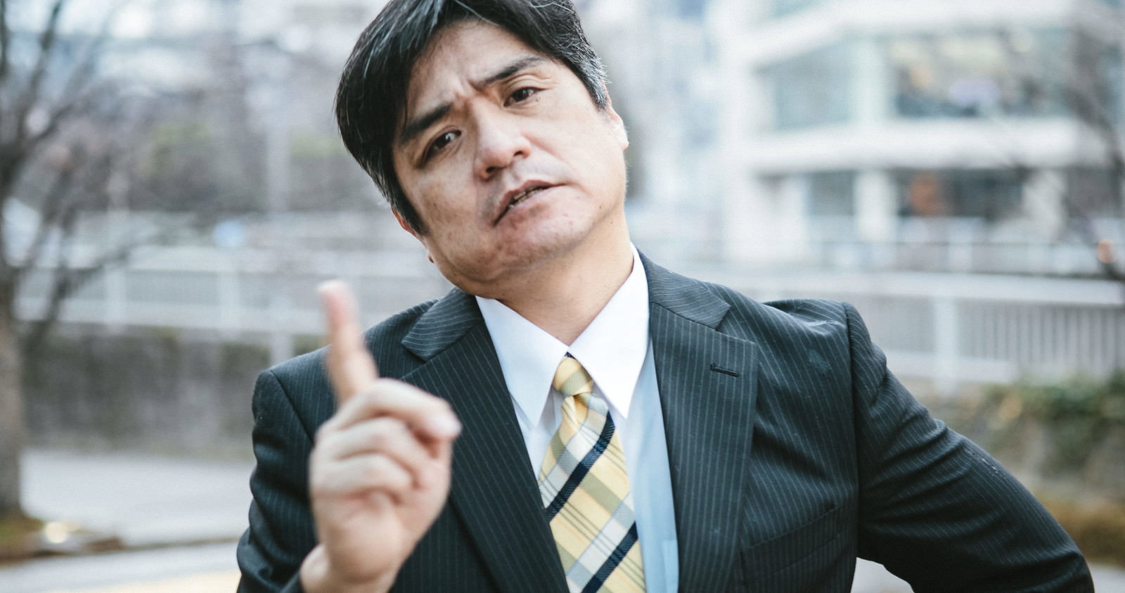黒いスーツを着た男性が不機嫌そうな表情で上を見上げている様子［モデル：よたか］