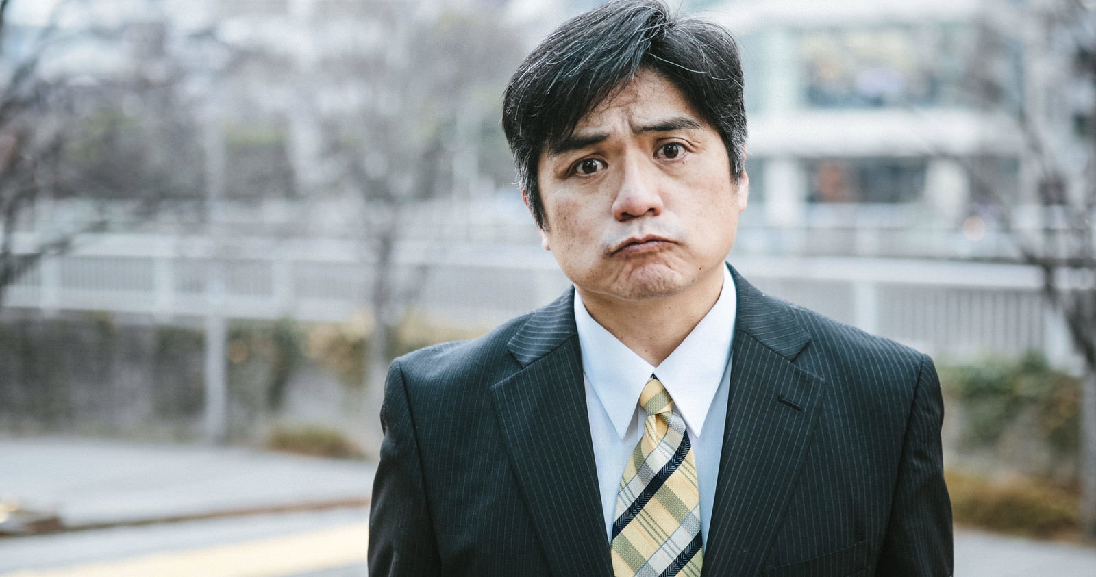 眉をひそめ不機嫌な表情の男性部長がスーツ姿で立っている［モデル：よたか］
