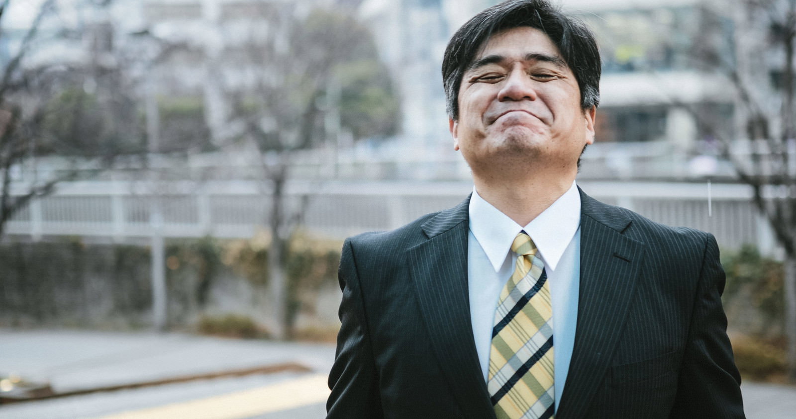 スーツと黄色チェック柄ネクタイを着た中年男性が、斜め上を見上げて口角を下げた表情で耐えている［モデル：よたか］
