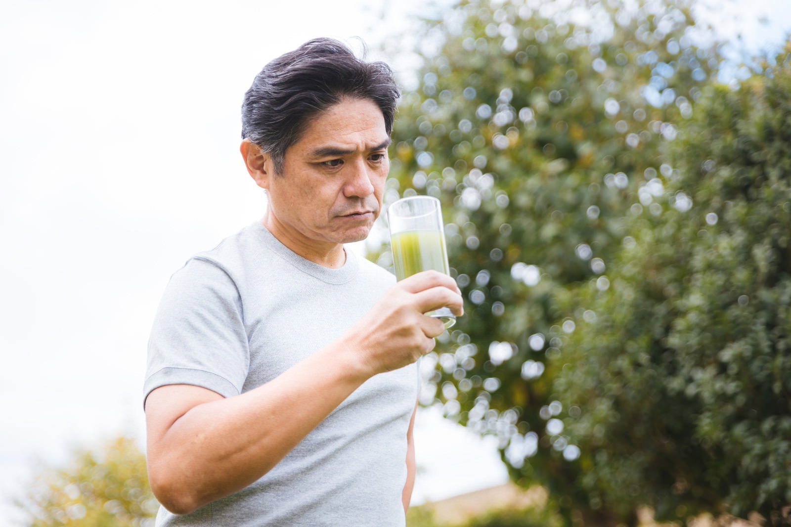 青汁入りのコップを手に持ち、屋外で飲んでいる中年男性［モデル：よたか］