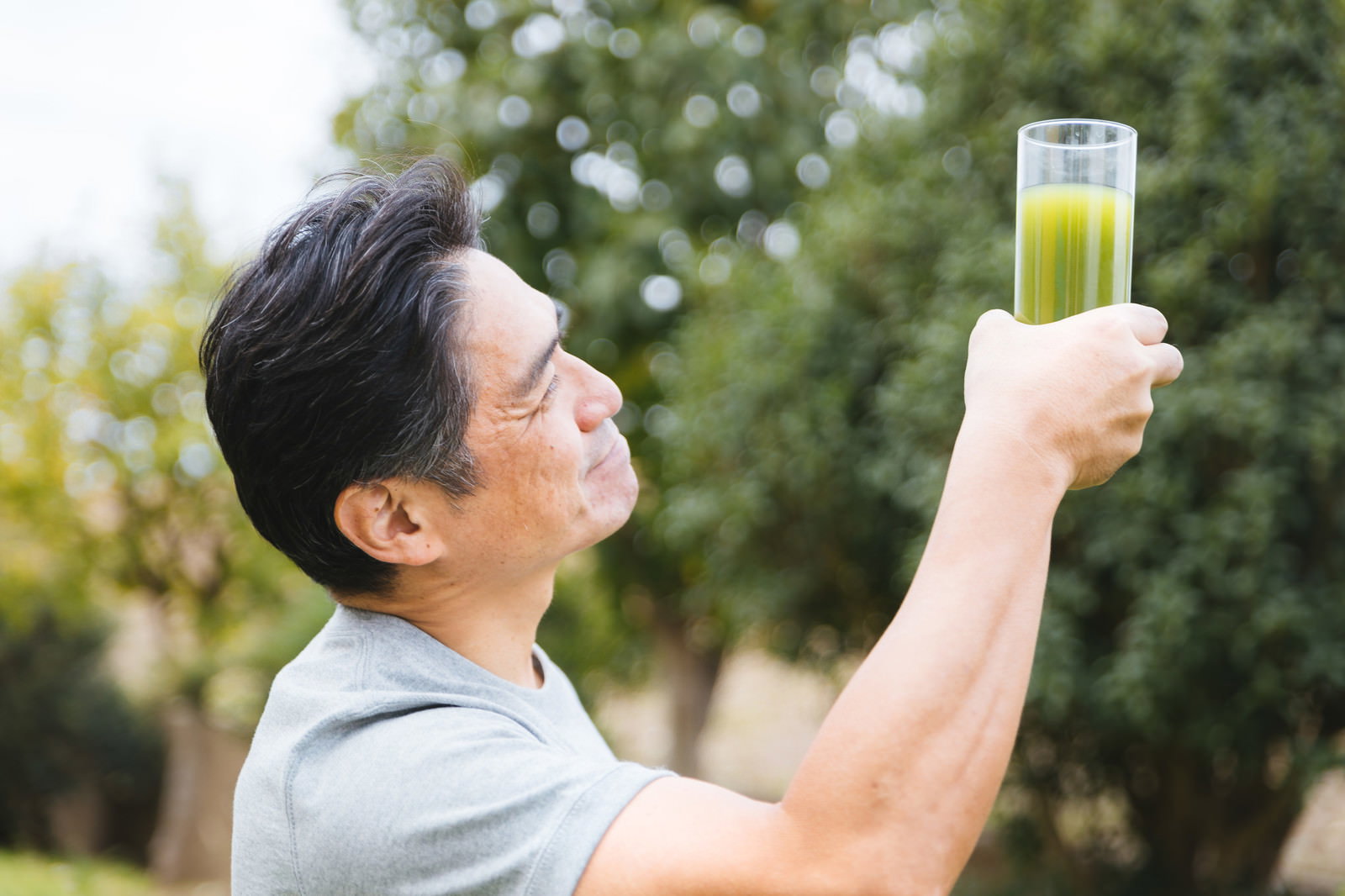 屋外で青汁入りグラスを持ち上を向いて飲む中年男性［モデル：よたか］