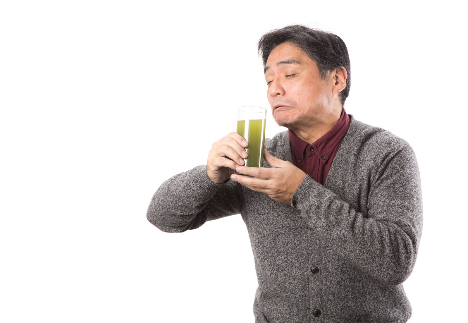 グレーのセーターを着た中年男性が青汁の入ったグラスを持って飲んでいる［モデル：よたか］