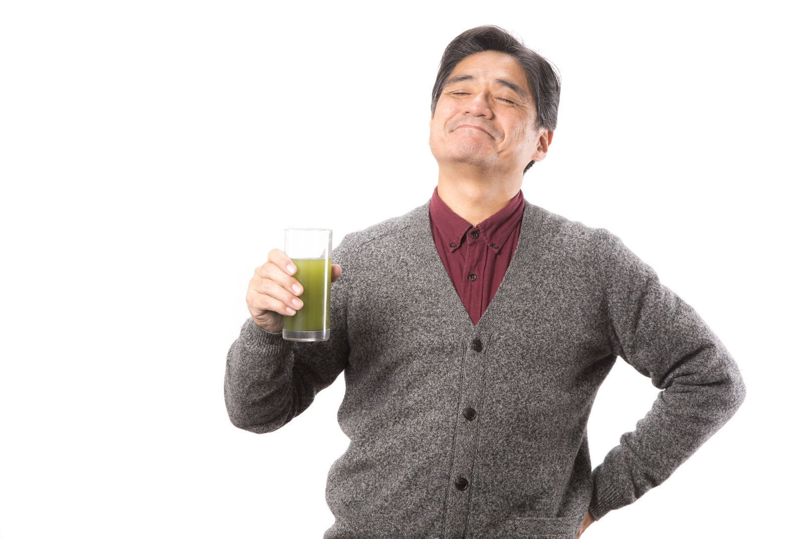 グレーのベストを着て青汁を飲む中年男性が腰に手を当てて立っている［モデル：よたか］