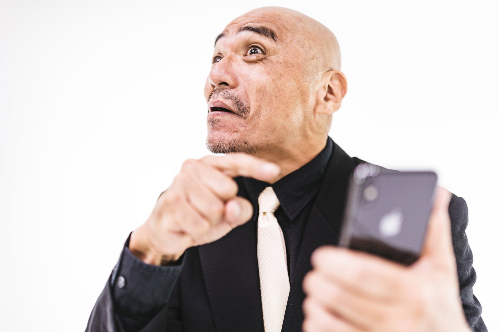黒いスーツ姿でスマートフォンを持ち、顎に手を触れながら上を見上げるスキンヘッドの中年男性［モデル：よたか］