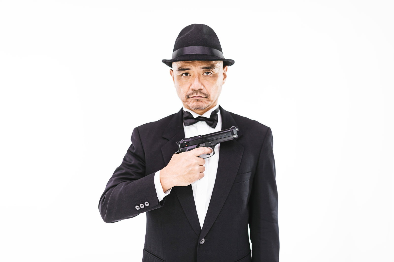 黒いスーツと帽子を着用し拳銃を構えるポーズをとる中年男性［モデル：よたか］