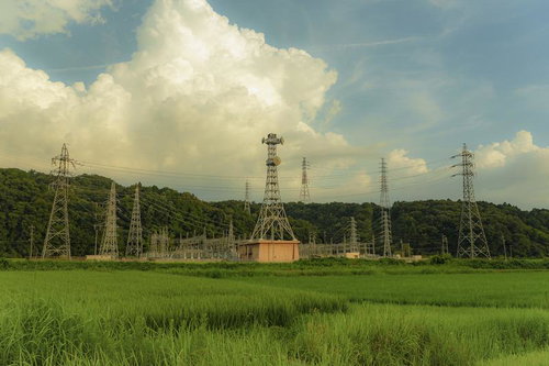 青空と積雲の下に立つ複数の送電鉄塔と緑の田園地帯