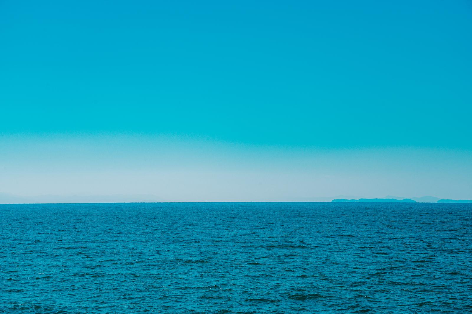 遠くに霞む島影と濃い青の海、淡い青空が広がる風景