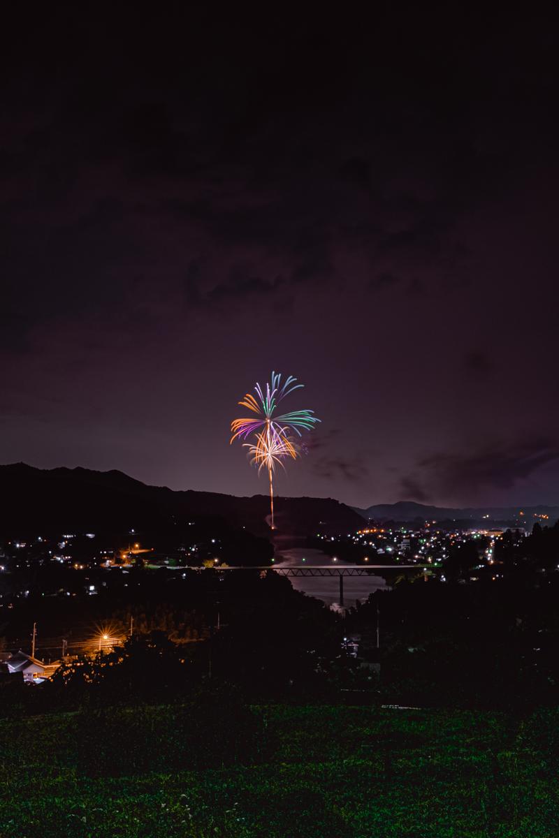 夜空で爆発する黄金色とカラフルな打ち上げ花火、下方に町の夜景と川が見える