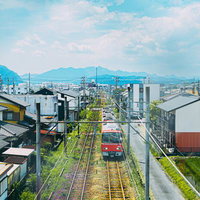 青空と山並みを背景に住宅街を走る赤いローカル電車の写真