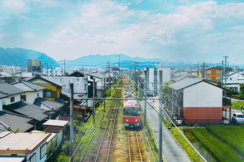 青空と山並みを背景に住宅街を走る赤いローカル電車
