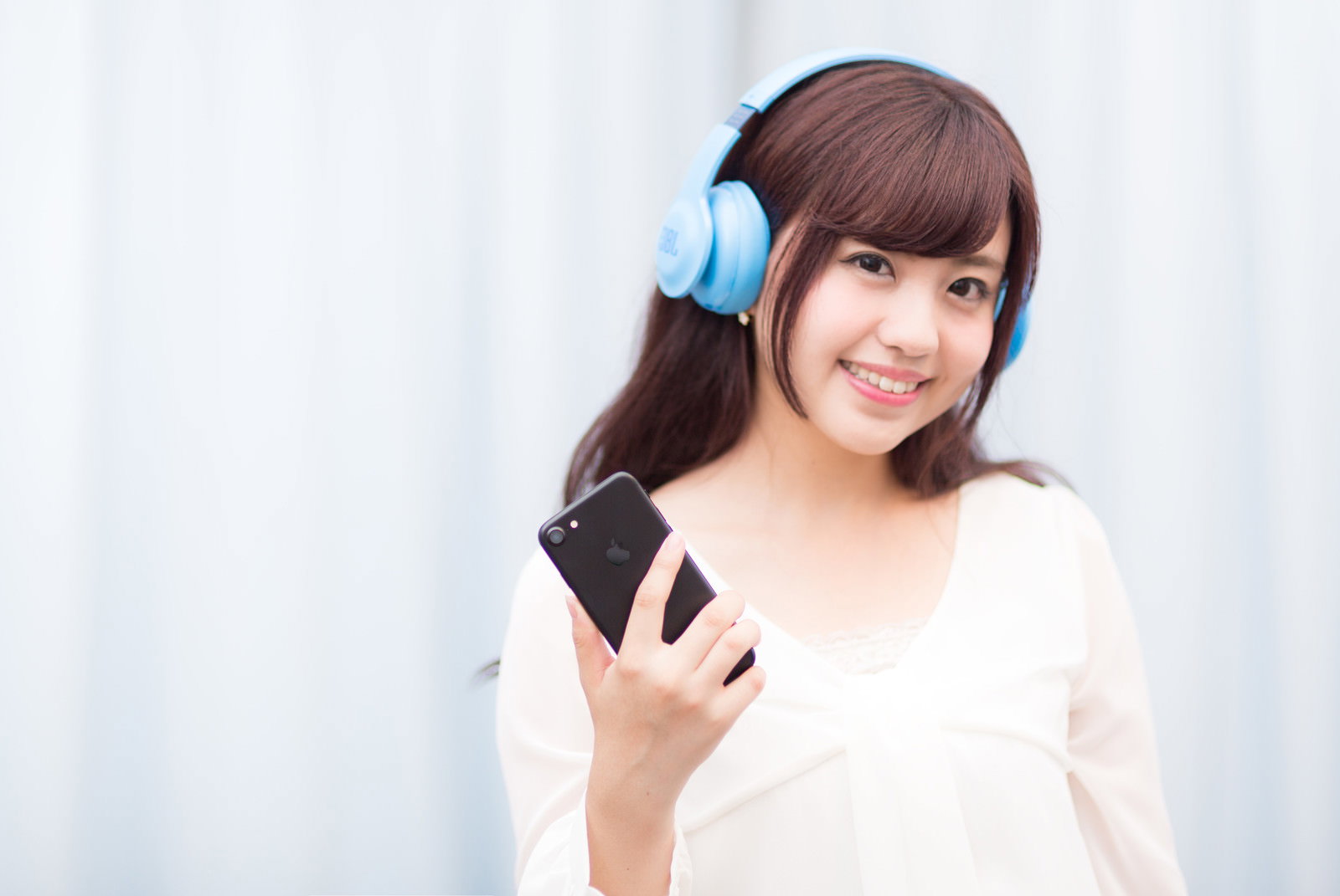 ブルーのヘッドフォンをつけてスマホを持つ笑顔の女性［モデル：河村友歌］