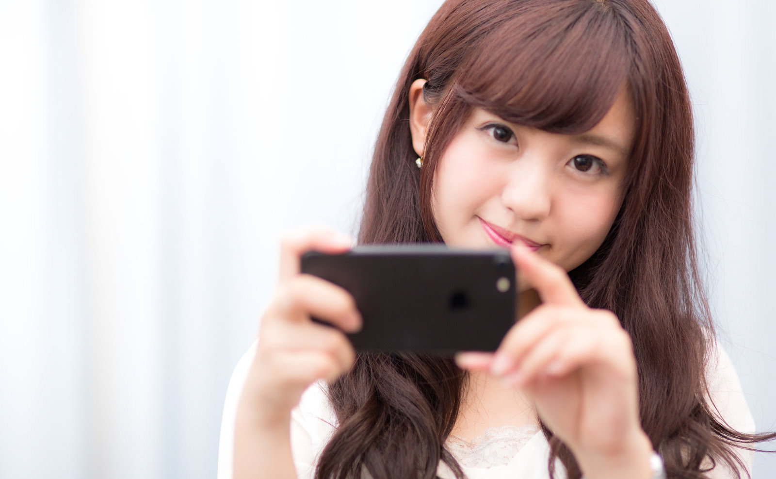 黒いスマートフォンを両手で構えて笑顔の女性［モデル：河村友歌］